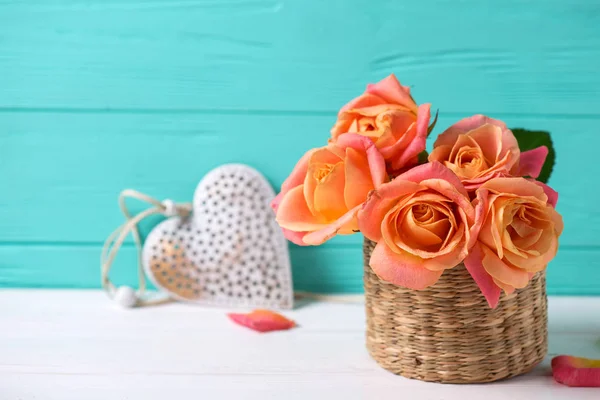 Fresh orange roses and heart on white wooden background against turquoise wall. Place for text. Floral still life.