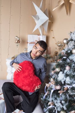 Genç adam büyük kırmızı kalbi tut ve Noel ağacı ve şöminenin yanındaki koltukta otur.