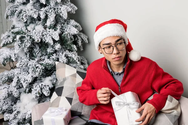 Fiatal férfi kalapban és pulóverben ül a kanapén a karácsonyfa mellett. — Stock Fotó