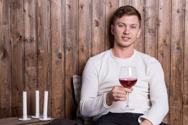 Genç yakışıklı bir adam bir bardak şarap ve kırmızı bir kalple tahta duvarın yanında oturuyor.
