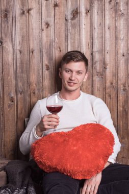Genç yakışıklı bir adam bir bardak şarap ve kırmızı bir kalple tahta duvarın yanında oturuyor.
