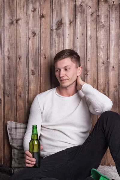 Joven hombre guapo sentado cerca de la pared de madera y beber cerveza — Foto de Stock