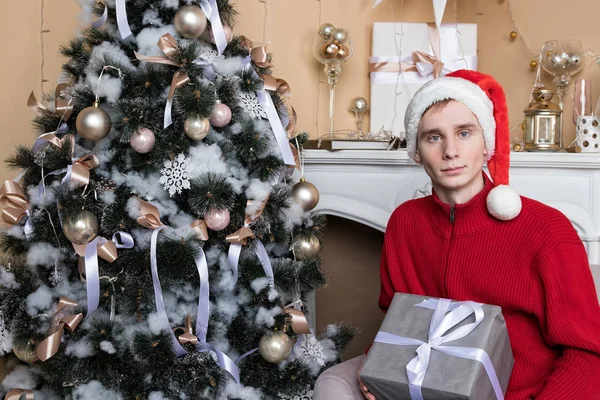 Fiatal férfi piros pulóverben, ajándék ül a kanapén közel karácsonyfa és kandalló — Stock Fotó