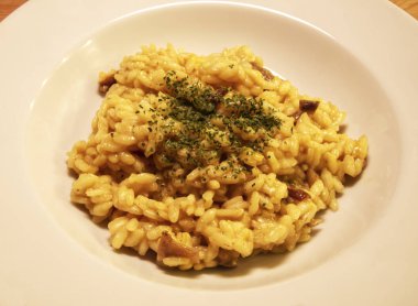 Geleneksel İtalyan pilavı, mantarlı, safranlı, maydanozlu, beyaz tabakta, ahşap bir masa.