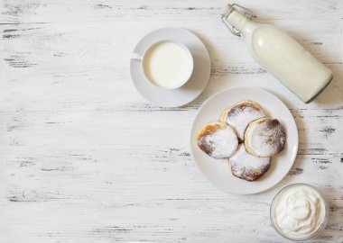 Süzme peynirli krep. Peynirli kekler. Syrniki veya sirniki. Sağlıklı kahvaltı. Beyaz tabaklar. Bir şişe süt. Bir bardak süt. Yoğurt veya ekşi krema. Beyaz arka plan. Üst Manzara. Metin için boşluk