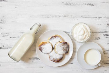 Süzme peynirli krep. Peynirli kekler. Syrniki veya sirniki. Sağlıklı kahvaltı. Beyaz tabaklar. Bir şişe süt. Bir bardak süt. Yoğurt veya ekşi krema. Beyaz arka plan. Üst Manzara. Metin için boşluk