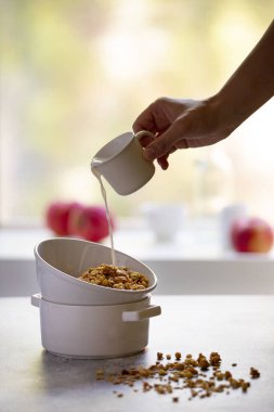 Beyaz kasede fındıklı ve kuru üzümlü granola. Sağlıklı kahvaltı pencerenin karşısındaki bir ışık masasında servis edilir. Süt bir bardaktan kaseye dökülüyor. Kadın eli süt döküyor. Dikey