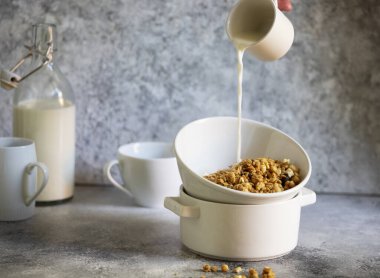Beyaz kasede fındıklı ve kuru üzümlü granola. Hafif bir masada servis edilen sağlıklı bir kahvaltı. Süt bir bardaktan kaseye dökülüyor. Kadın eli süt döküyor. Hafif nötr tonlar