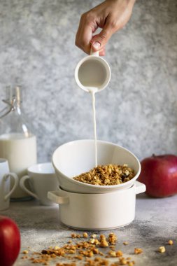 Beyaz kasede fındıklı ve kuru üzümlü granola. Hafif bir masada servis edilen sağlıklı bir kahvaltı. Süt bir bardaktan kaseye dökülüyor. Kadın eli süt döküyor. Hafif nötr tonlar