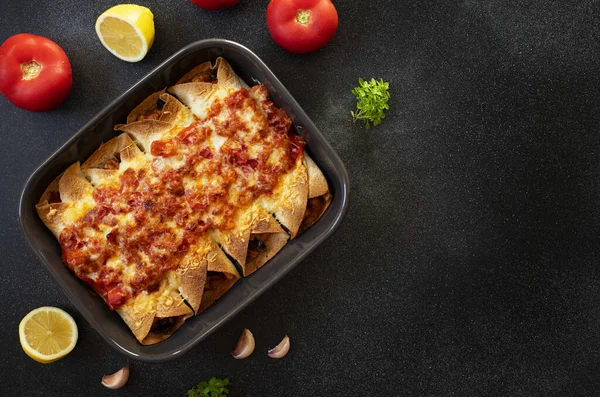 Mexican enchiladas with chicken, vegetables, corn, beans, tomato sauce and cheese. Served in baking tray. Mexican food. Latin American cuisine. Black background, top view, copy space