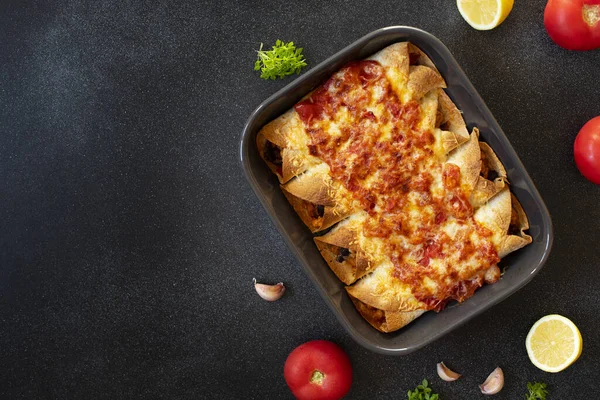 Mexican enchiladas with chicken, vegetables, corn, beans, tomato sauce and cheese. Served in baking tray. Mexican food. Latin American cuisine. Black background, top view, copy space — Stock Image Mexican Enchiladas Chicken Vegetables Corn Beans Tomato Sauce Cheese Served Stock Photo