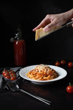 Domates soslu ve parmesanlı makarna. Siyah arka planda spagetti olan beyaz tabak. Makarnada parmesan peyniri rendeleyen eller. Geleneksel İtalyan yemekleri. Alçak tuş, dikey, kopyalama alanı