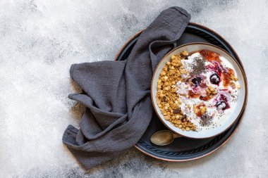 Fındıklı granola, Yunan yoğurdu, organik reçel ve chia tohumları beyaz bir kase ve bakır tepside servis edilir. Sağlıklı kahvaltı, sağlıklı beslenme konsepti. Gri arkaplan, üst görünüm, kopyalama alanı