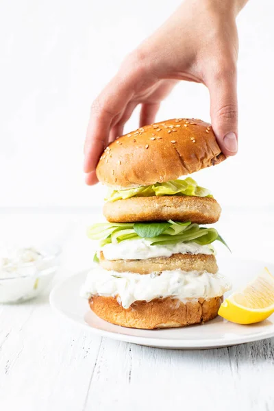 Delicious healthy crispy fish burger with greek yogurt-based sauce with lettuce and cucumber. Female hand putting a bun on a burger. White background, close-up, selective focus, copy-space