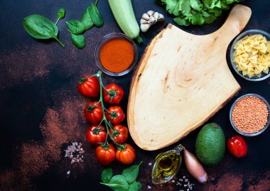 Çeşitli çiğ sebzeler, zeytin yağı, mercimek, baharat ve makarna ile çevrili boş ahşap kesme tahtası. Vejetaryen, vejetaryen yemeği, sağlıklı beslenme konsepti. Rustik arkaplan, üst görünüm, kopyalama alanı