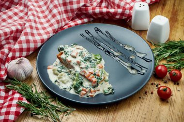 Krema soslu sebzeli ızgara somon bifteği, ahşap arka planda siyah tabak.