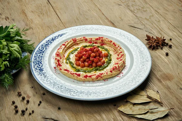 Hummus with red spices, chickpeas and herbs on a ceramic plate on a wooden background. Arabic or Israeli cuisine. Close up, horizontal. Snack