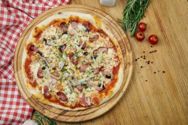 Tütsülenmiş sosisli pizza, soğan, mısır, jambon ve eritilmiş peynirli ahşap tepsi. Tahta bir masanın üzerindeki malzemelerle kompozisyon halinde pizza.