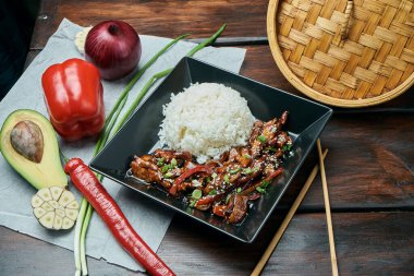 Klasik bir Asya yemeği. Ahşap bir arka planda siyah tabakta teriyaki kızarmış domuz tavası ile pilav. Kapat.