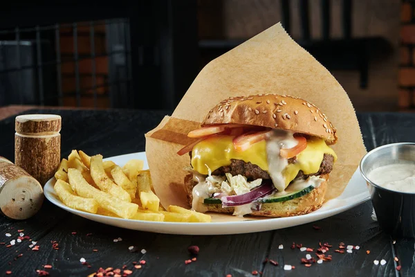 Appetizing and juicy burger with a large beef patty, melted cheddar cheese, fresh tomatoes, cucumber, onions and cabbage on a white plate with french fries.