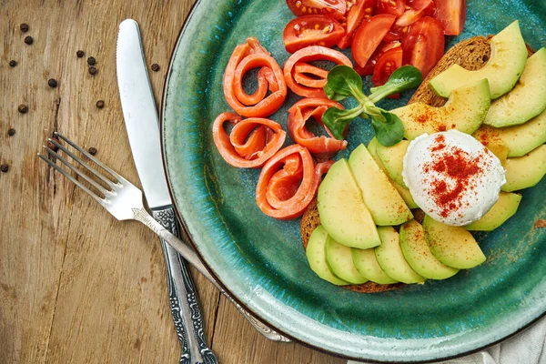 Trending food - avocado toast with poached egg on rye bread with sidedish salmon, tomato in a ceramic plate on a wooden background. Healthy breakfast meal