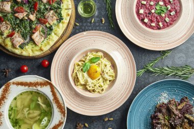Koyu arka planda çeşitli İtalyan yemekleri: Carbonara makarna, somon pizza, pancar pilavı, ev yapımı tavuk suyu ve salata. Üst manzara düz yiyecek yatıyordu.