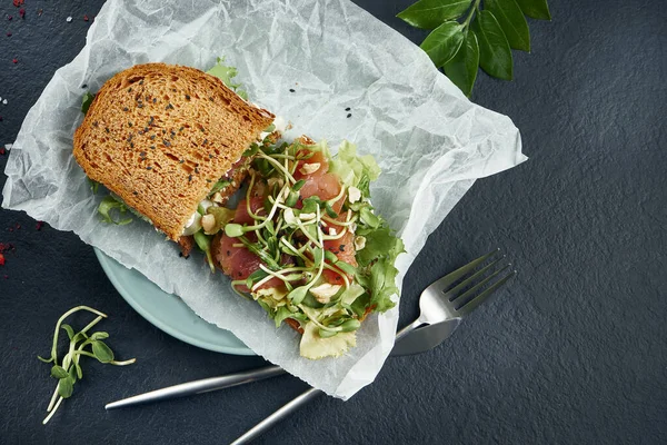 Trendy street snack. Tasty sandwich toast with salmon and microgreen on craft paper on a black background. Top view. Flay lay food with copy space. Seafood