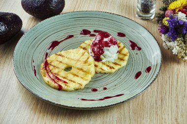 Izgara dilimlenmiş ananas, bir kepçe beyaz vanilyalı dondurma ve gri bir tabağa kırmızı reçel dökülmüş. Yemekten sonra lezzetli bir tatlı. Kapatın. Ahşap arkaplan