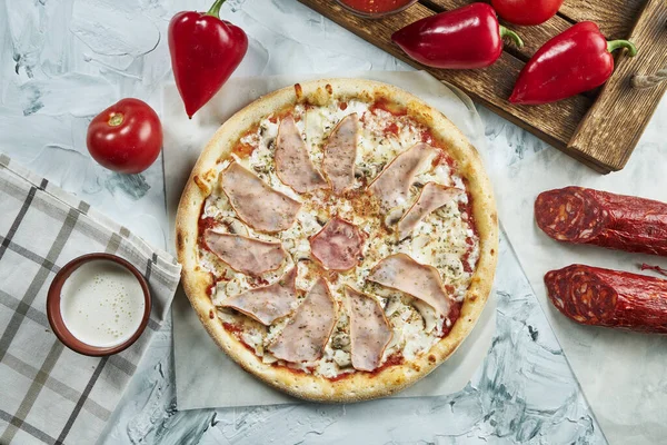 Homemade oven-baked meat pizza with tomatoes, cheese, ham, red sauce. Composition with crispy pizza, tomatoes on a light background. Copy space. Top view