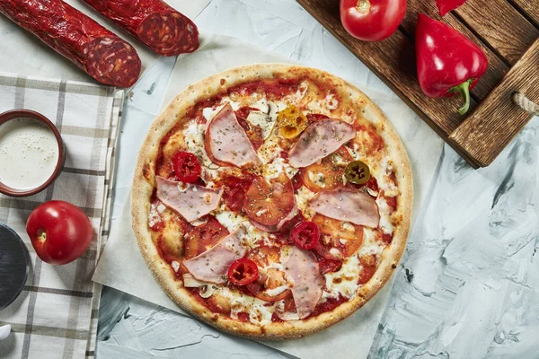 Homemade oven-baked meat pizza with tomatoes, cheese, ham, red sauce. Composition with crispy pizza, tomatoes on a light background. Copy space. Top view