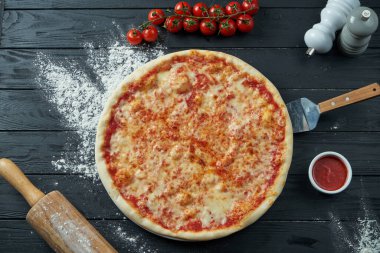 Fırında margarita pizza, domatesli ve erimiş peynir, siyah ahşap arka planda kırmızı sos ve malzemeler. Üst görünüm