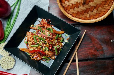 Udon eriştesi, bebek mısırı, dolmalık biber, soğan, havuç ve tavada kızartılmış dana eti ve ahşap arka planda siyah tabakta teriyaki sosu. Lezzetli Asya sokak yemekleri