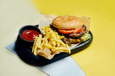 İştah açıcı Amerikan hamburgeri, sığır eti, domates, marul ve patates kızartması renkli bir arka planda siyah tepside. Lezzetli fast food.