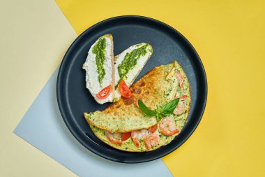 Karides, domates ve fesleğenli omlet krem peynirli tost ve siyah bir tabakta pesto..