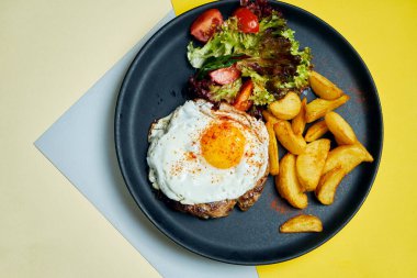 Biftek, sahanda yumurta, yanında salata ve siyah bir tabakta kızarmış patates. Öğle yemeği için iştah açıcı yemek.
