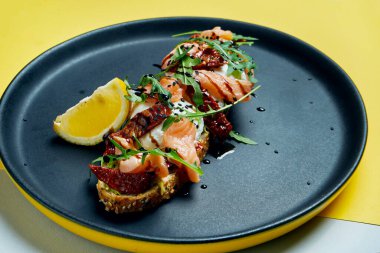 Çavdar ekmeğinin üstünde roka, haşlanmış yumurta, somon balığı ve siyah bir tabakta kurutulmuş domatesle iştah açıcı tostu yakından izleyin. Kahvaltı hazır.