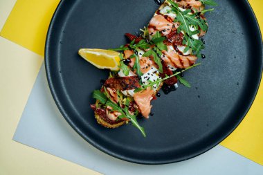 Çavdar ekmeğinin üstünde roka, haşlanmış yumurta, somon balığı ve siyah bir tabakta kurutulmuş domatesle iştah açıcı tostu yakından izleyin. Kahvaltı hazır.