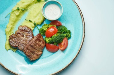 Brokolili ızgara sığır eti madalyonu, vişneli domates ve gri arka planda izole edilmiş mavi bir tabakta yeşil püre. Kapat.