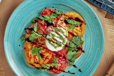 Farklı domates türlerinden taze salata ve ahşap arka planda mavi bir tabakta pesto ve balzamik soslu burrata peyniri servis ediliyor. Restoran masası ayarı