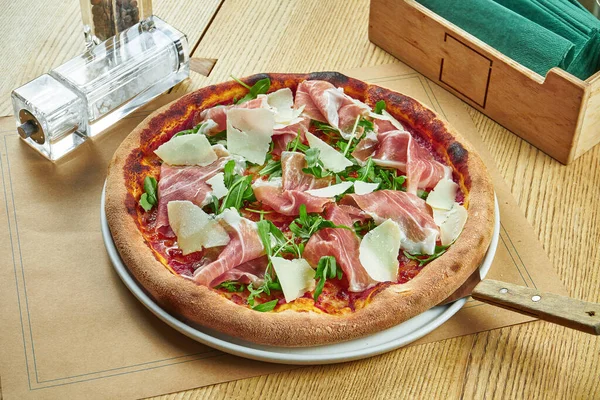 Appetizing baked pizza with prosciutto di parma, parmesan and arugula with crispy crust on a wooden background. Restaurant table setting.Close up