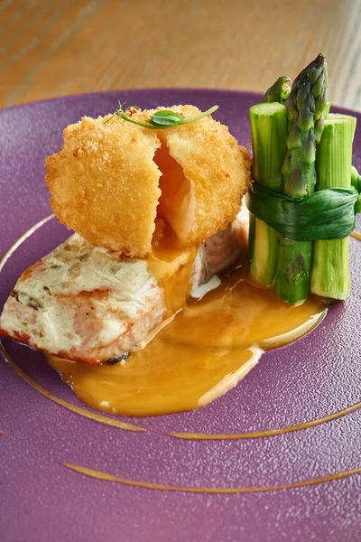 An appetizing restaurant dish is chicken croquet with a flowing yolk on a salmon steak with a side dish of asparagus on a purple plate. Wood background. Selective focus.