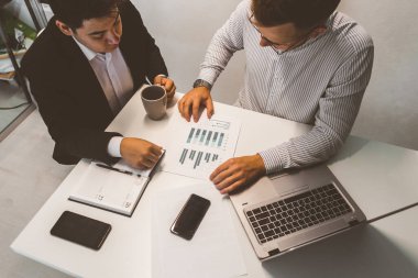 Masada oturan genç bir ofis çalışanı, bilgisayar kullanıyor. İki iş adamı konuşuyor.
