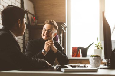 Masada oturan genç bir ofis çalışanı, bilgisayar kullanıyor. İki iş adamı konuşuyor.