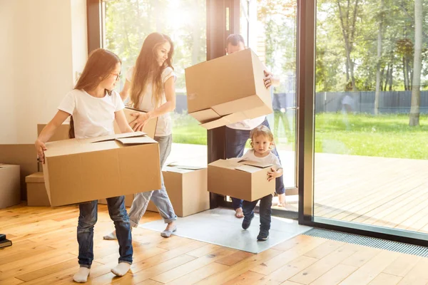 Feliz familia joven, padres hija e hijo, desempacando cajas y mudarse a un nuevo hogar. niños divertidos corren con cajas — Foto de Stock