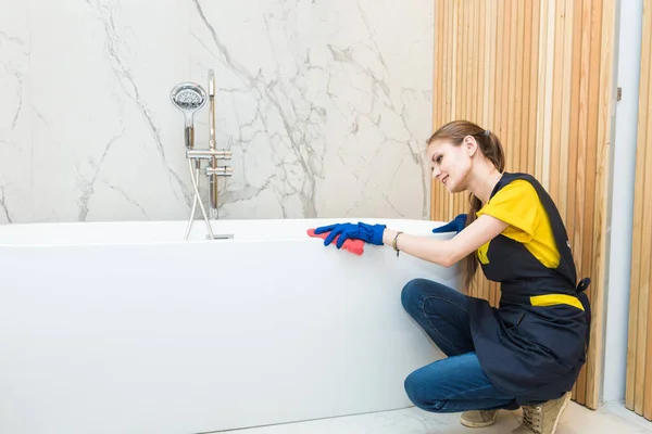 Servicio de limpieza profesional. mujer en uniforme y guantes esponja lava la plomería en el baño. Bañera y lavabo —  Fotos de Stock