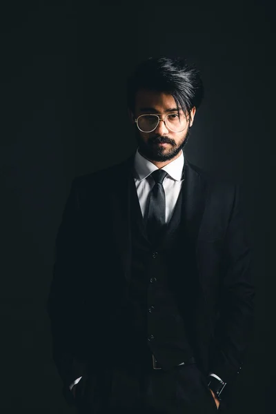 Retrato de un joven guapo y exitoso indio con un clásico traje de negocios negro de tres piezas sobre un fondo oscuro, mantenga presionadas las gafas —  Fotos de Stock