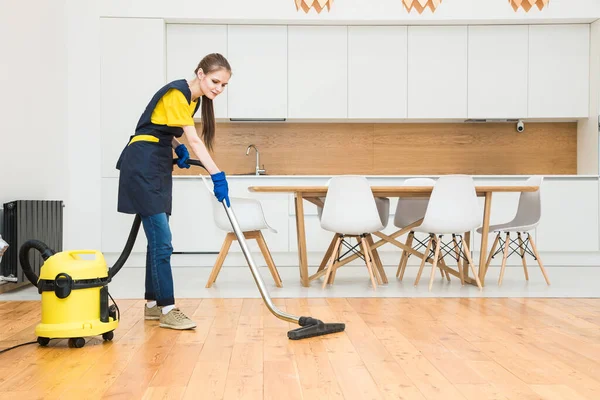 Servicio de limpieza profesional. mujer en uniforme y guantes hace la limpieza en una cabaña. el trabajador aspira el suelo con equipo profesional — Foto de Stock