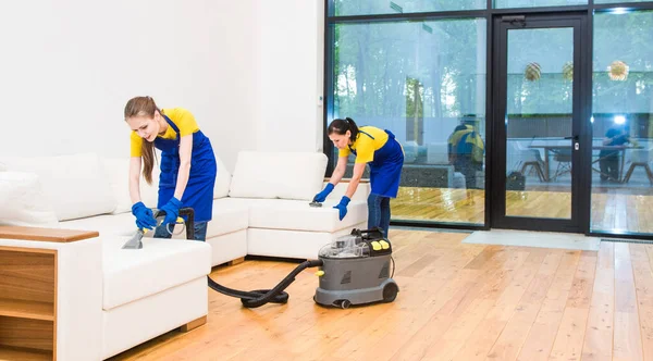 Profesyonel temizlik servisi. Önlüklü, işçi üniformalı iki kadın. Önlüklü, işçi üniformalı iki kadın, özel evin temizliğini bölüşüyorlar. Yıkama, süpürme, kanepeyi temizleme. — Stok fotoğraf