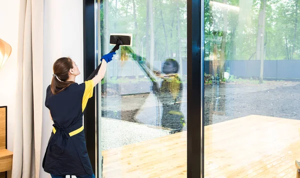 Servizio di pulizia professionale. donna in uniforme e guanti spugna lava finestre panoramiche nel cottage. lavaggio mocio a vapore e raschietto — Foto Stock