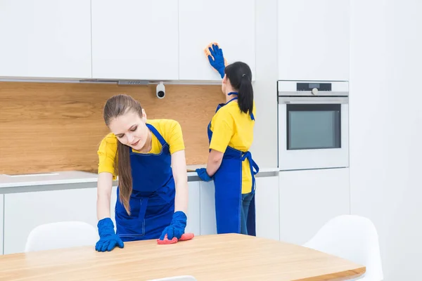 Professionell städning. Två kvinnor i arbetsuniform, i förkläden, dela städning av köket i ett privat hus, stuga. Tvätta kylskåpet, knacka, handfat. Tvättgolv. — Stockfoto